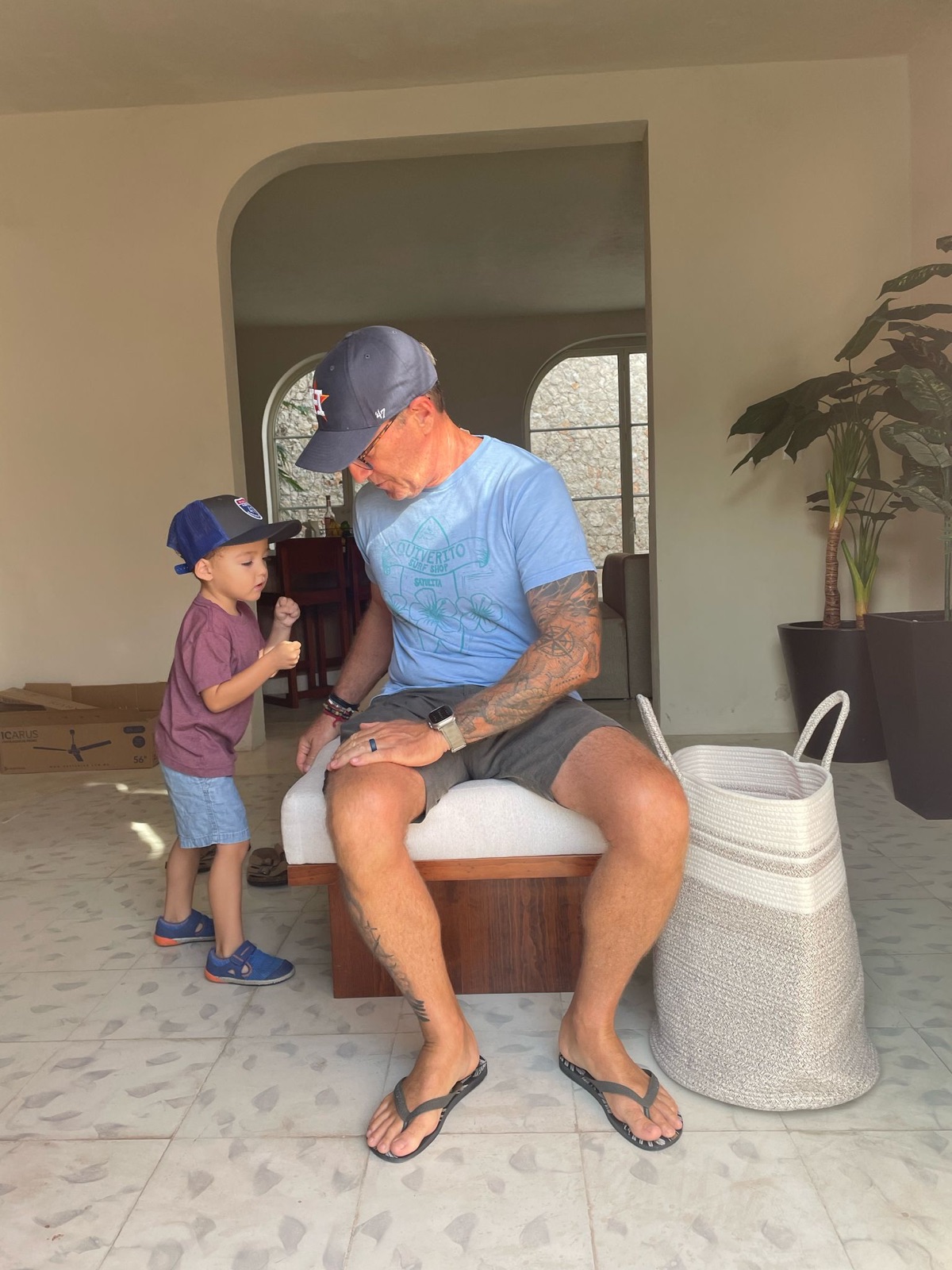 Rob seated at home, looking down at his son mid fist-bump beside him — an ordinary, unposed moment.