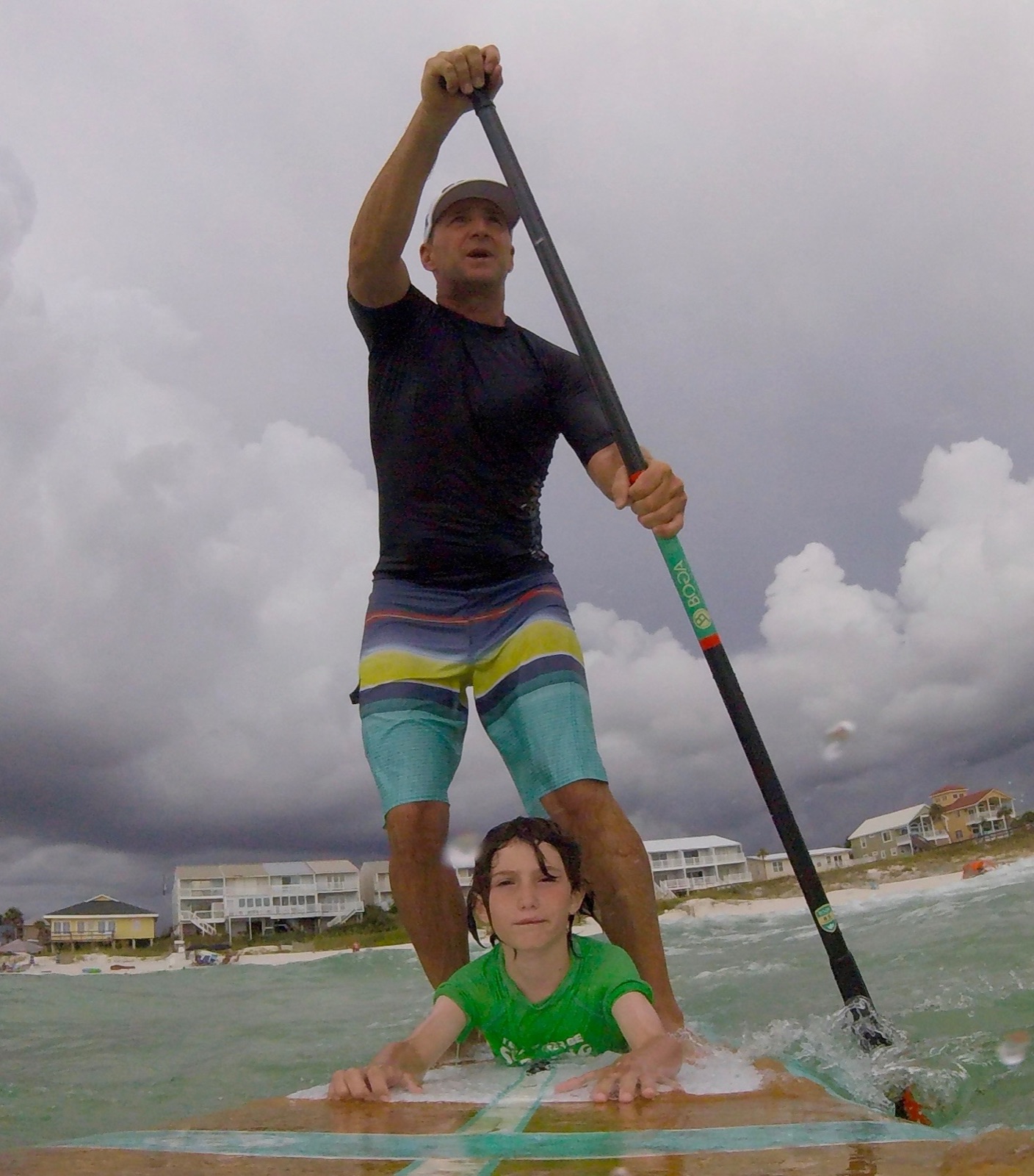 Rob standing on a paddleboard in choppy, storm-lit water, a child lying on the board in front of him, beach houses in the distance.