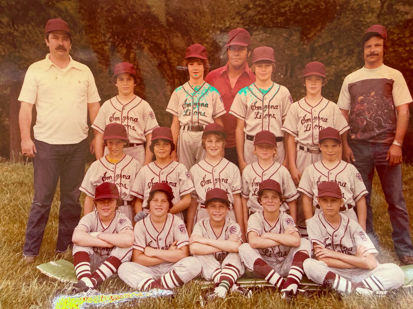 A faded color team photo of the Smyrna Lions youth baseball team on a grassy field: fourteen boys in maroon caps and “Smyrna Lions” uniforms with striped socks, posed in three rows—the front row sitting cross-armed on the grass—with three coaches standing at the back and sides. Rob is in the middle row, dead center.